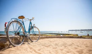 Vieux vélo bleu garé sur une plage bretonne ensoleillée avec vue sur la mer et les bateaux.
